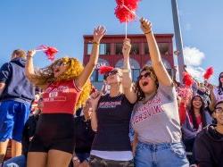 Three female students in a large crowd at homecoming cheering for the Montclair football team